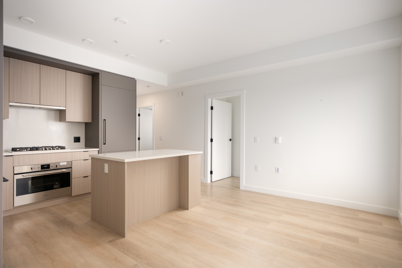 Modern kitchen with light wood cabinetry, an island, built-in appliances, and an open layout leading to adjoining rooms with light-colored walls and flooring.