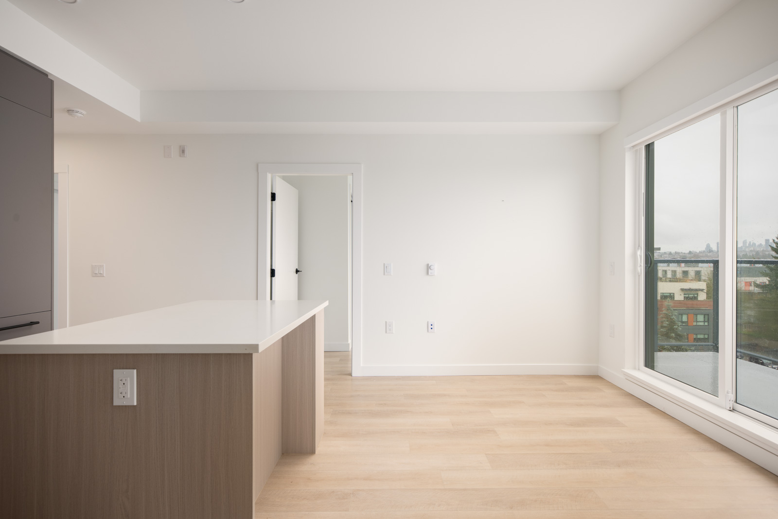A modern, unfurnished apartment room with light wood flooring, white walls, a kitchen island, and large windows overlooking a cityscape.