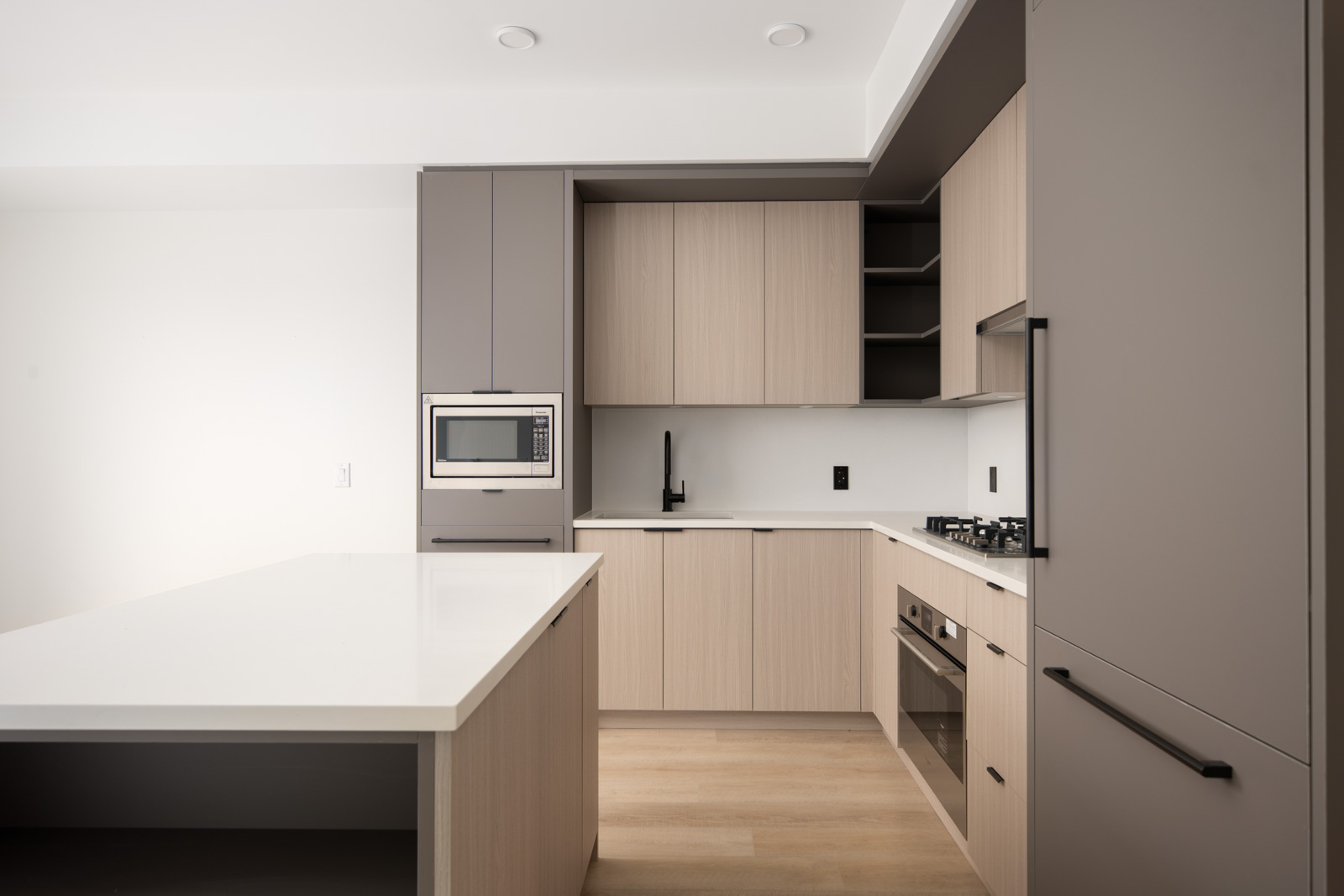 Modern kitchen with light wood cabinets, built-in microwave, oven, stovetop, large island with white countertop, and minimalist fixtures.