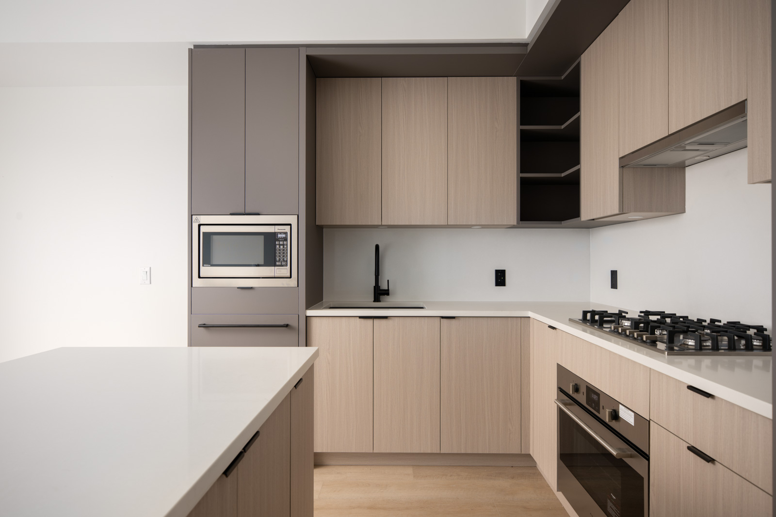 Modern kitchen with light wood cabinets, built-in microwave, black sink faucet, gas stove, oven, white countertops, and open shelving.