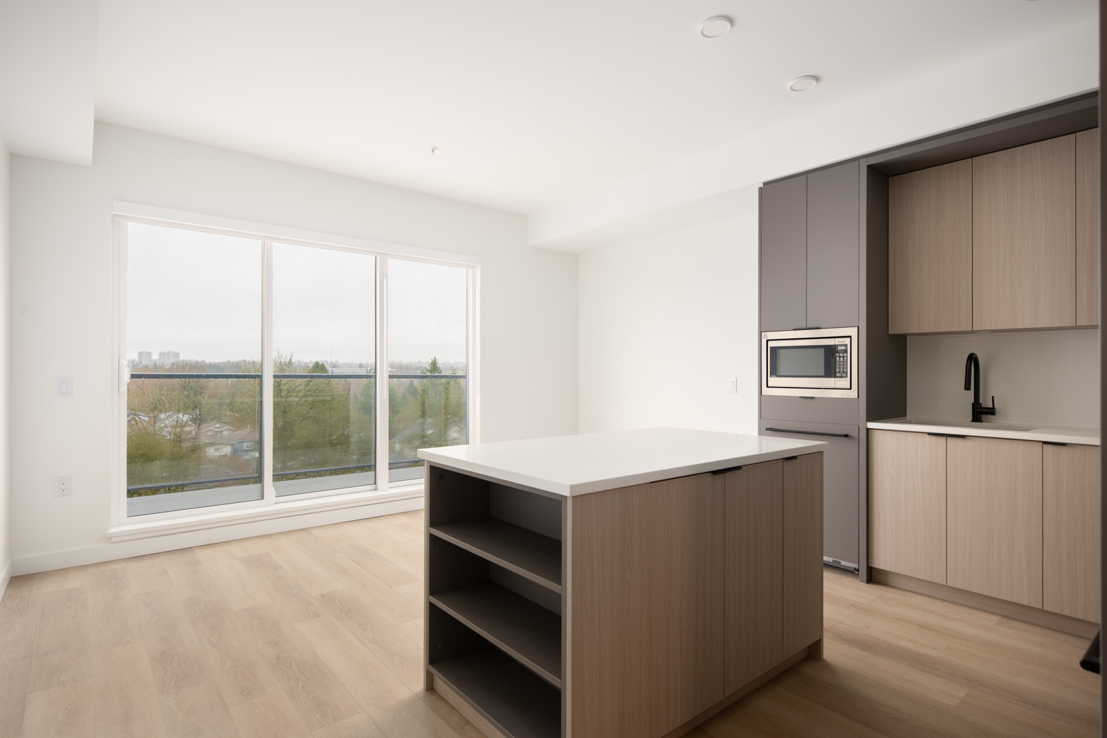 Modern, unfurnished kitchen with light wood floors, an island with open shelves, built-in microwave, and large windows overlooking trees and a cityscape.