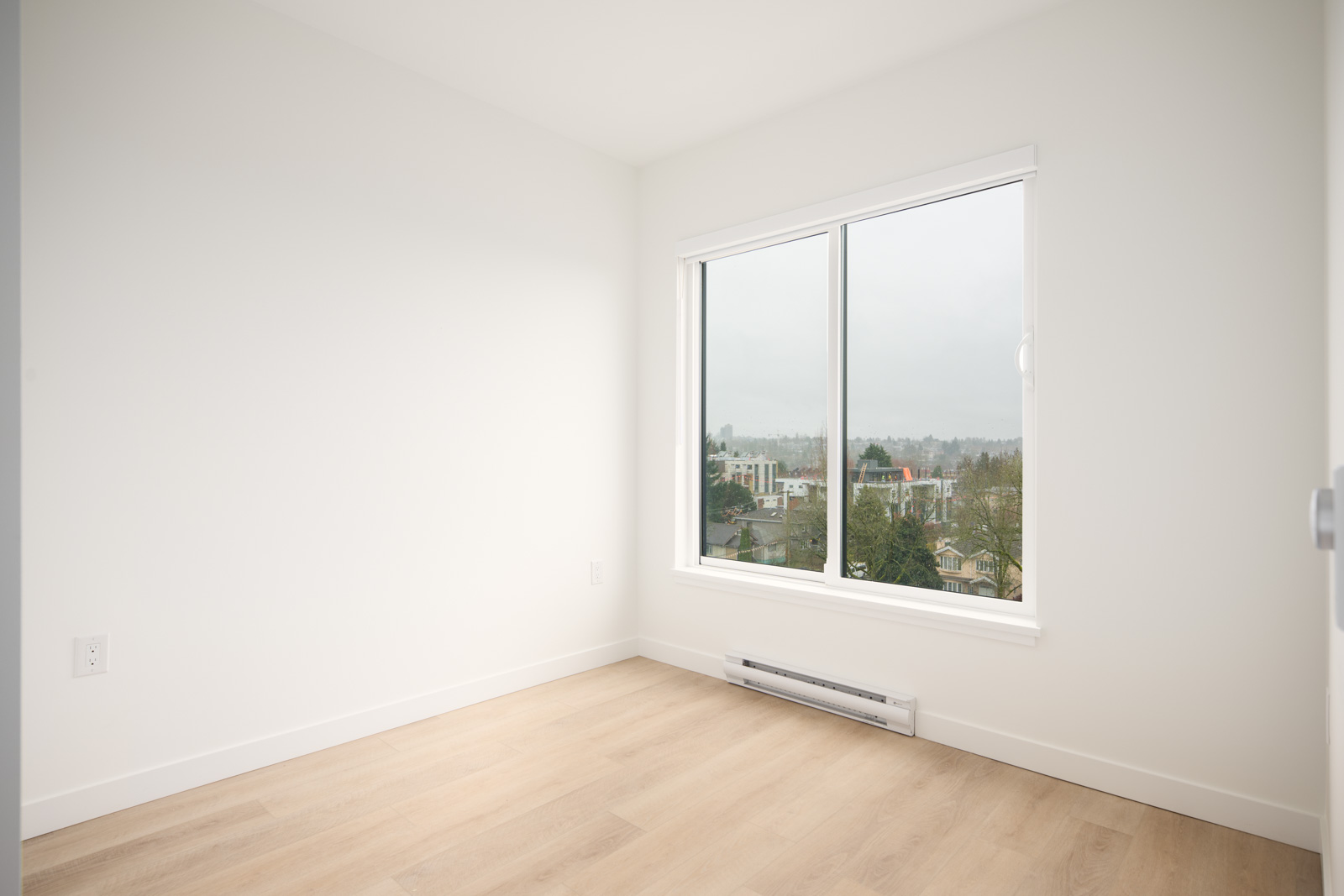 Empty, unfurnished room with light wood flooring, white walls, and a large window overlooking buildings and trees on a cloudy day.