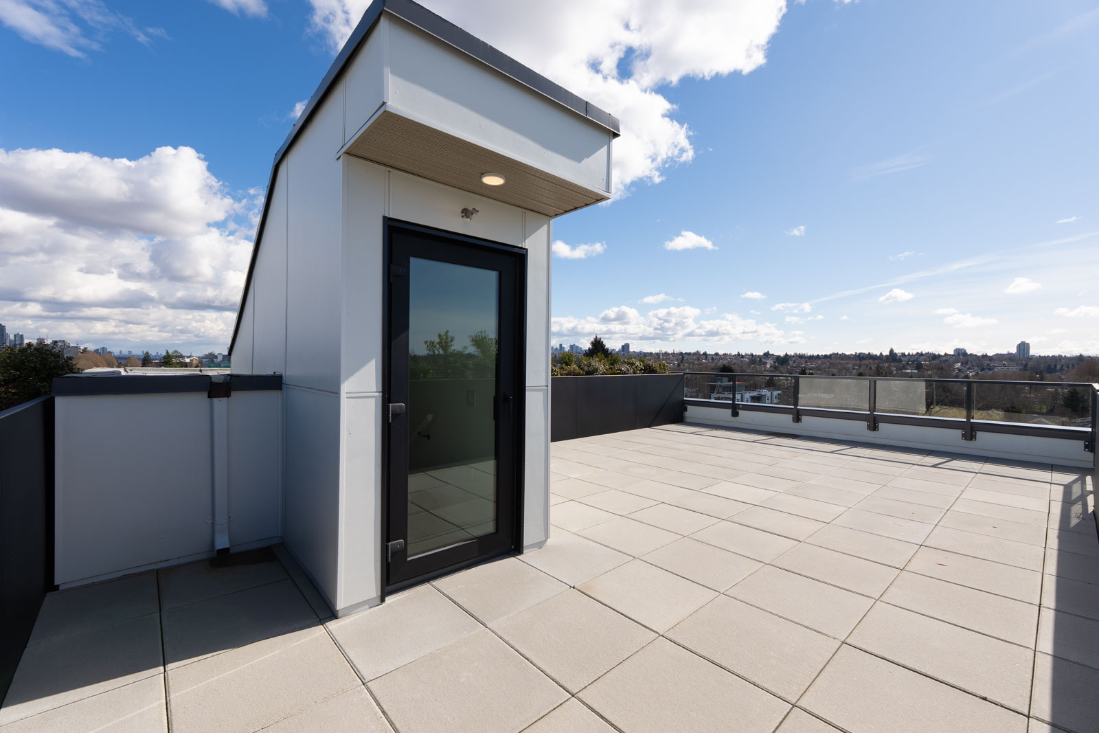Spacious rooftop terrace with tiled flooring, glass railing, and a small structure with a black-framed door under a partly cloudy sky.