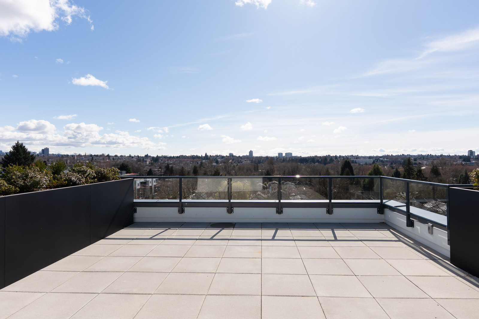 Rooftop patio with glass railing overlooking a cityscape and trees under a blue sky with scattered clouds.
