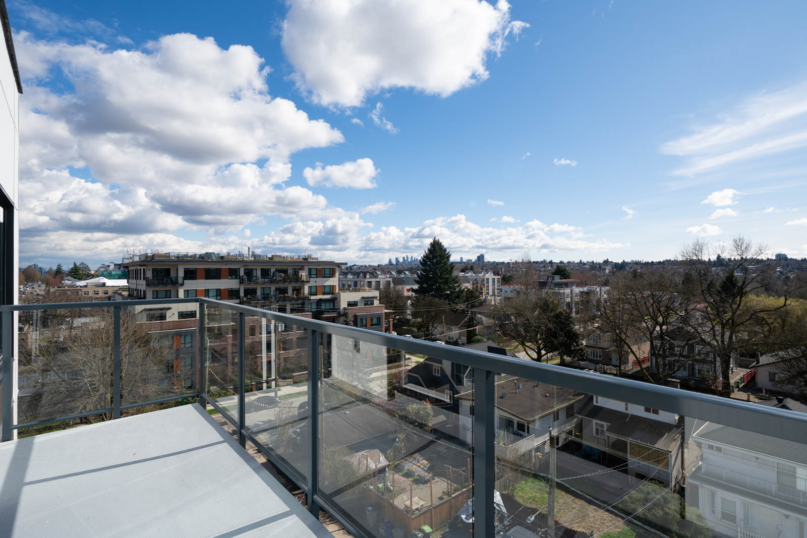 View from a balcony with glass railing overlooking residential buildings, trees, and a distant city skyline under a partly cloudy sky.