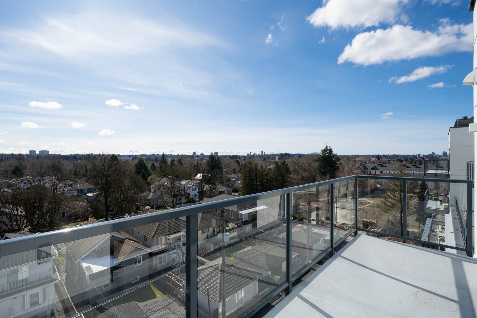 View from a high-rise balcony with glass railing overlooking a residential neighborhood, trees, and a distant city skyline under a blue sky with scattered clouds.