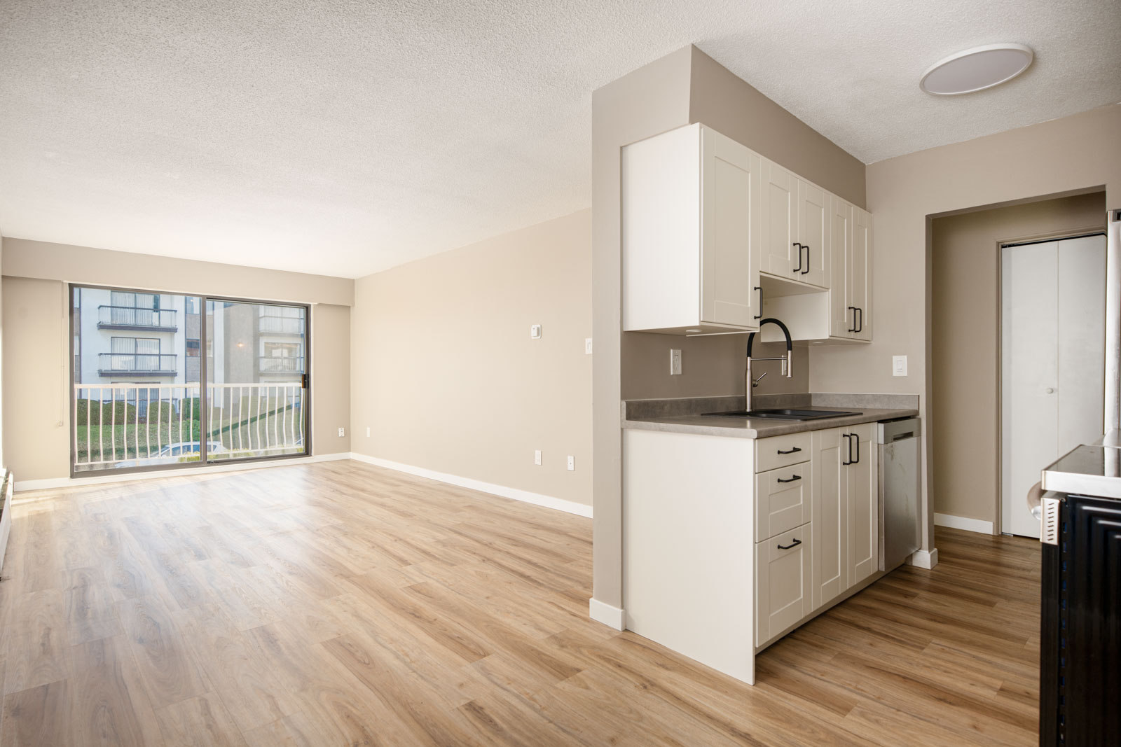 Unfurnished apartment with wood-look flooring, white kitchen cabinets, and sliding glass doors leading to a balcony with an exterior building view.