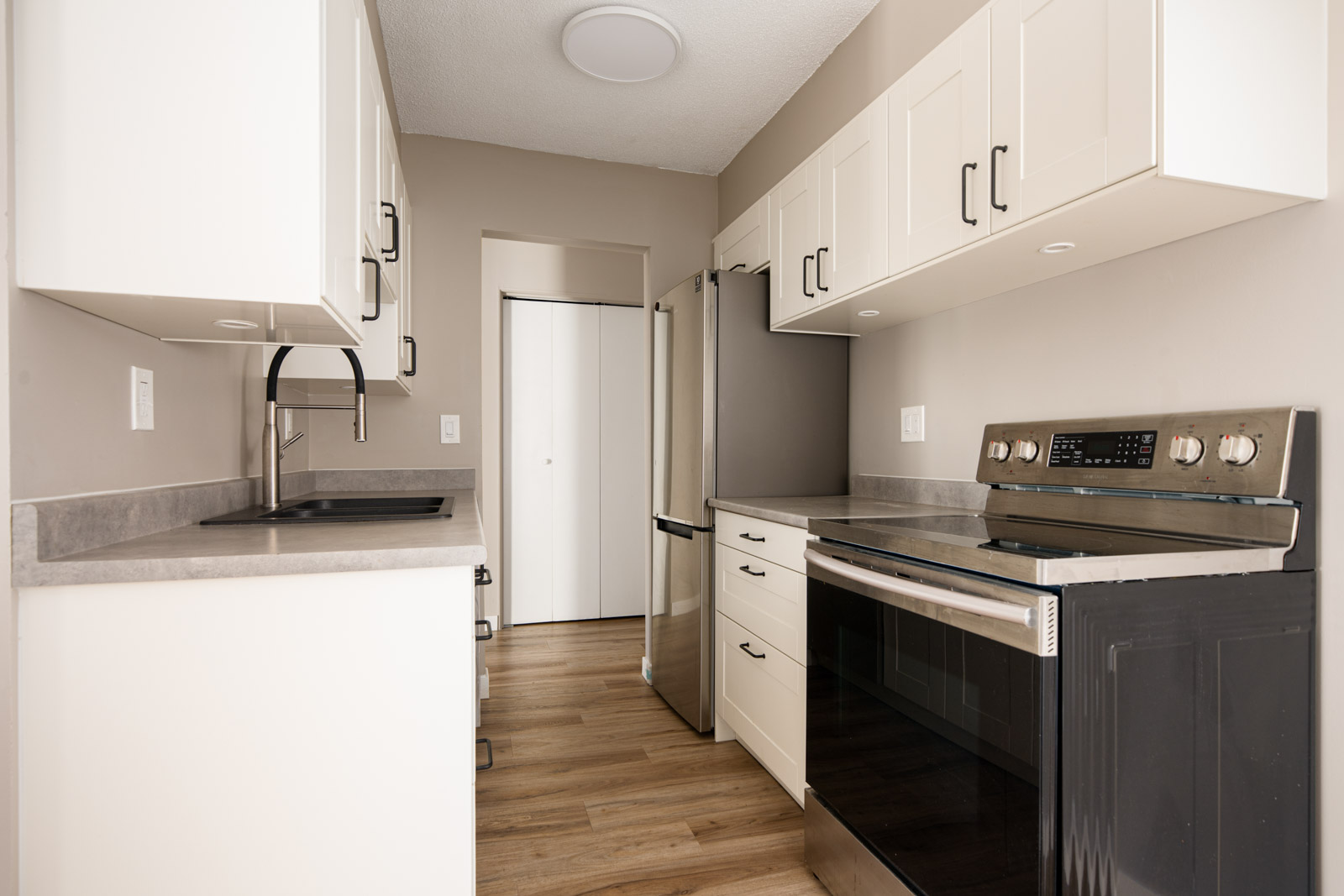 Modern galley kitchen with white cabinets, stainless steel appliances, gray countertops, and light wood flooring.