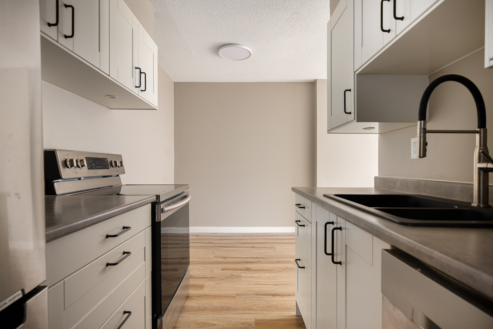 Modern galley kitchen with white cabinets, stainless steel appliances, black faucet, light wood flooring, and neutral-colored walls.