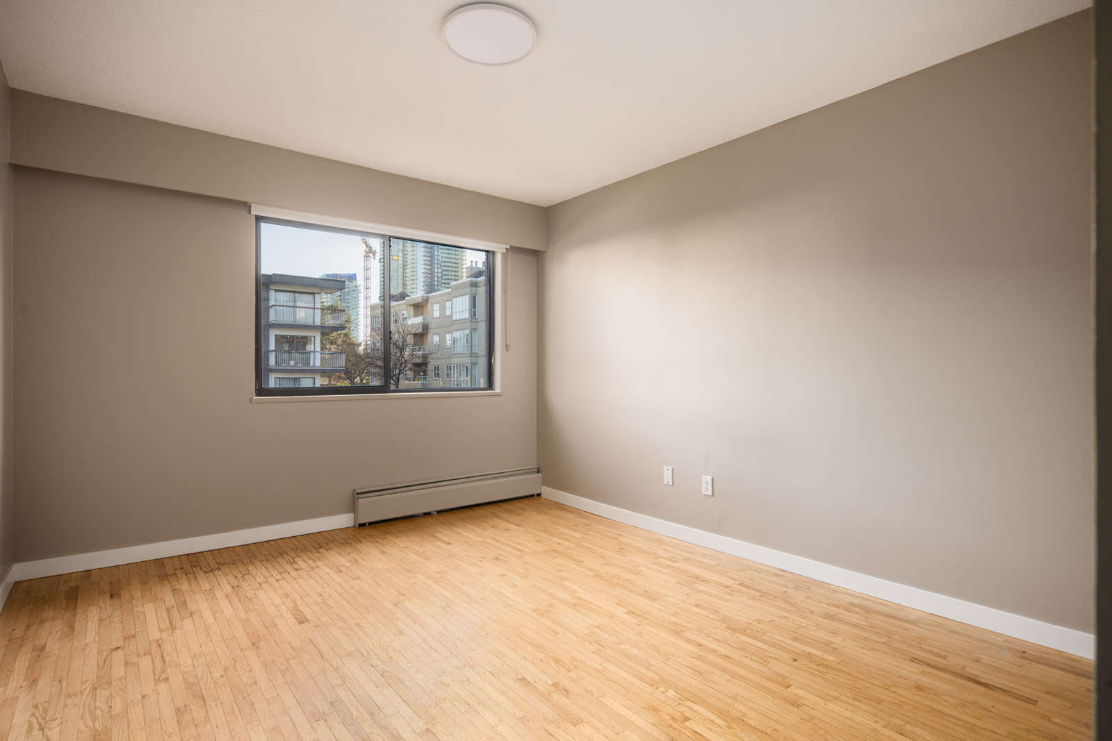 Empty room with light brown hardwood floor, beige walls, a large window, and baseboard heater beneath the window. Urban buildings are visible outside.