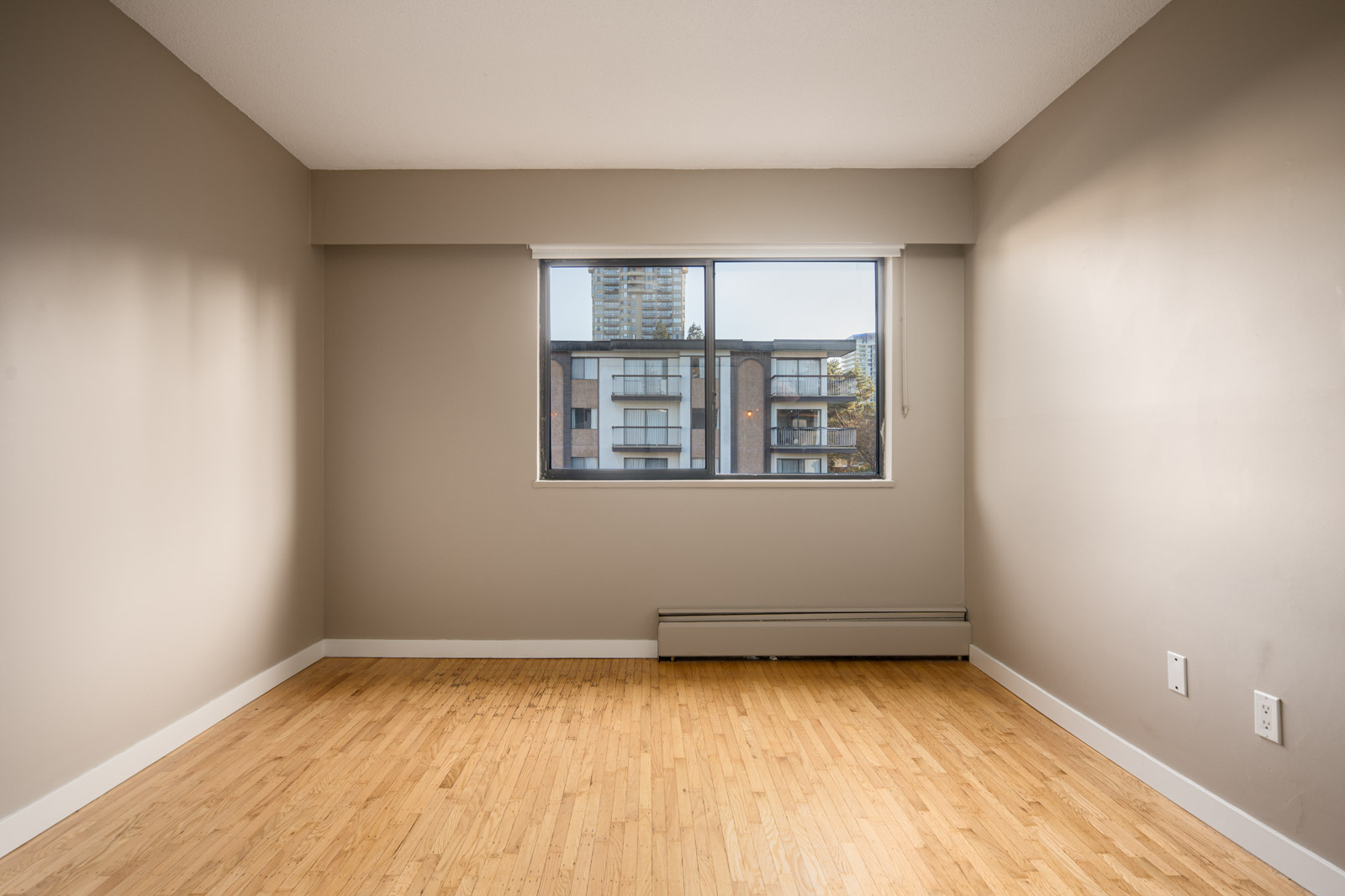 Empty room with beige walls, light wood flooring, and a large window overlooking neighboring apartment buildings.