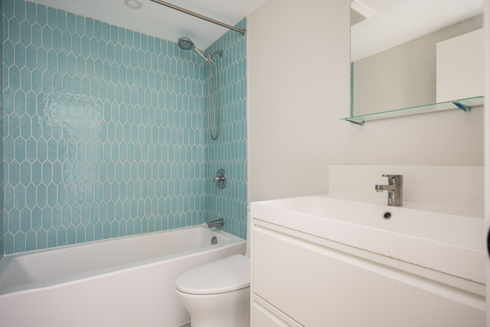 Modern bathroom with a blue geometric tile shower, white bathtub, toilet, and a white vanity with a mirror above it.