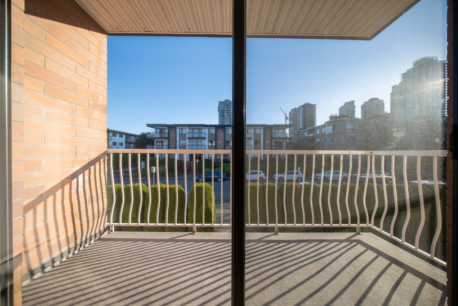 View from a small apartment balcony with a metal railing, overlooking a parking lot and several mid-rise buildings under a clear blue sky.