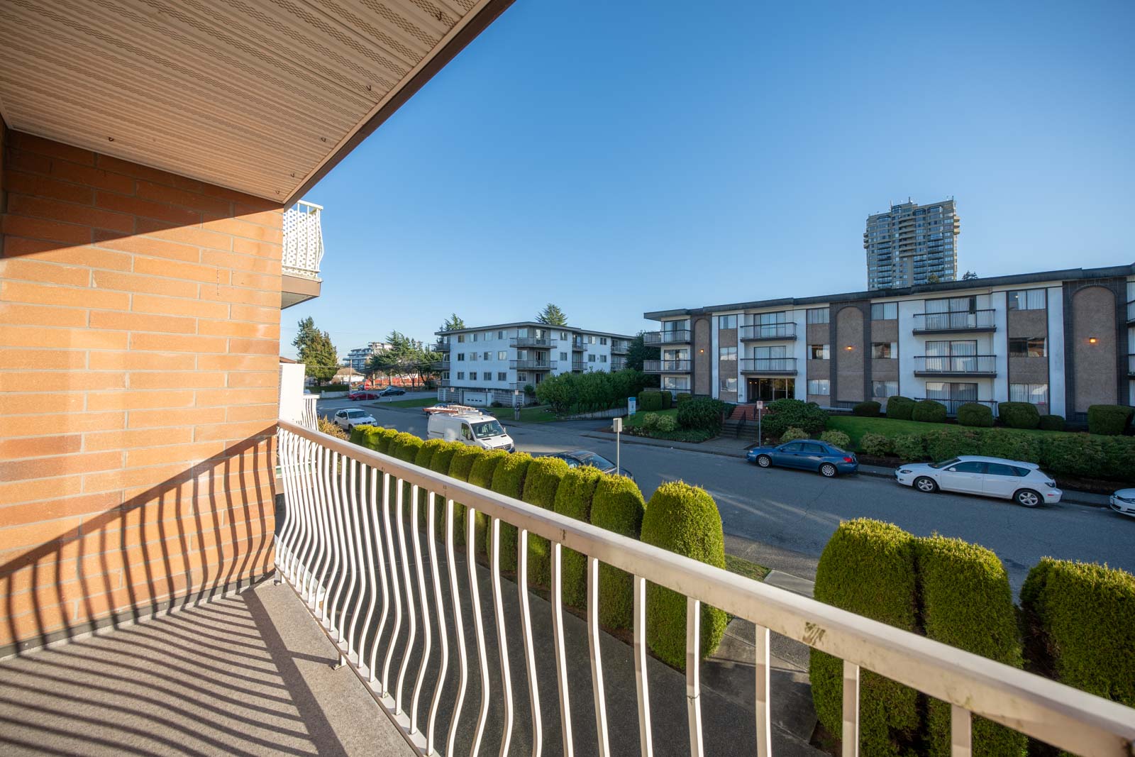 View from a balcony overlooking a quiet residential street with parked cars, green hedges, low-rise apartments, and a tall building in the background under a clear blue sky.