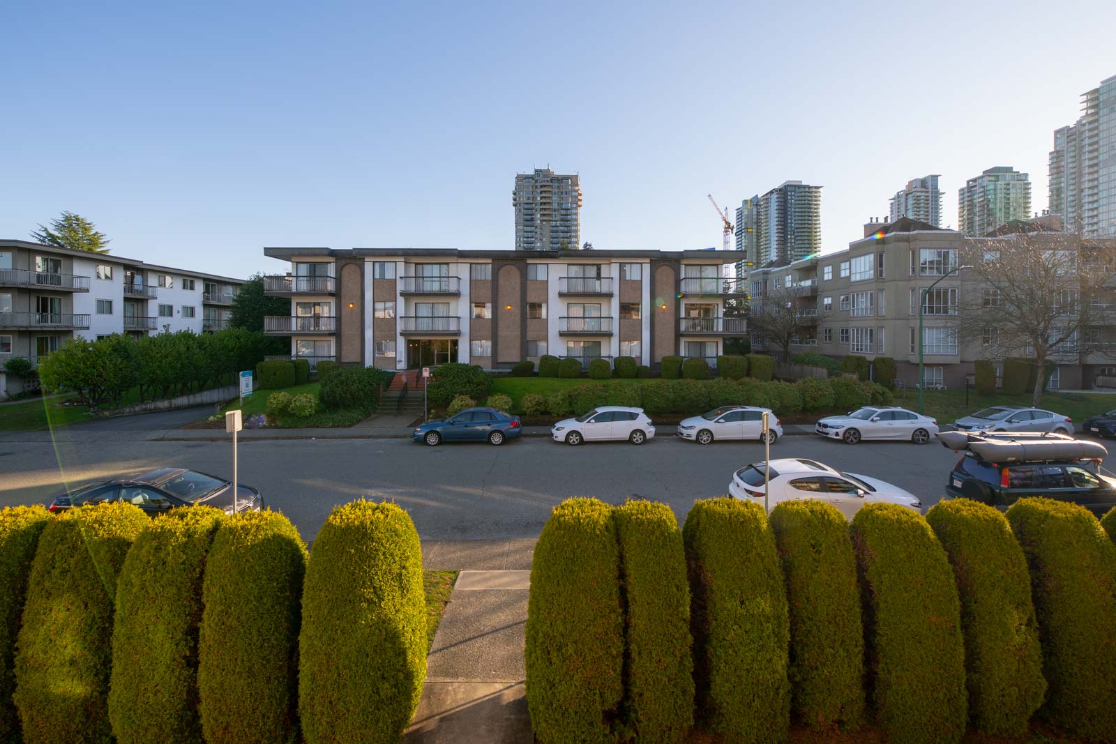 A mid-century apartment building with balconies, parked cars in front, and high-rise buildings in the background on a sunny day.
