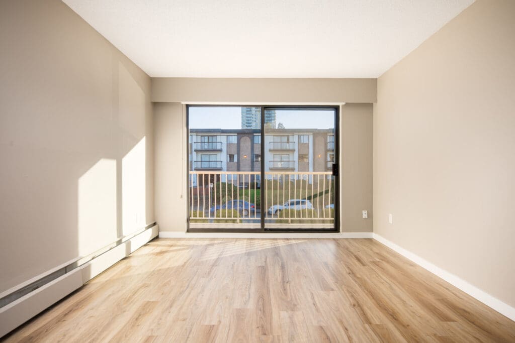 Empty room with light wood flooring, beige walls, and a large window with a sliding glass door leading to a balcony overlooking other apartment buildings.