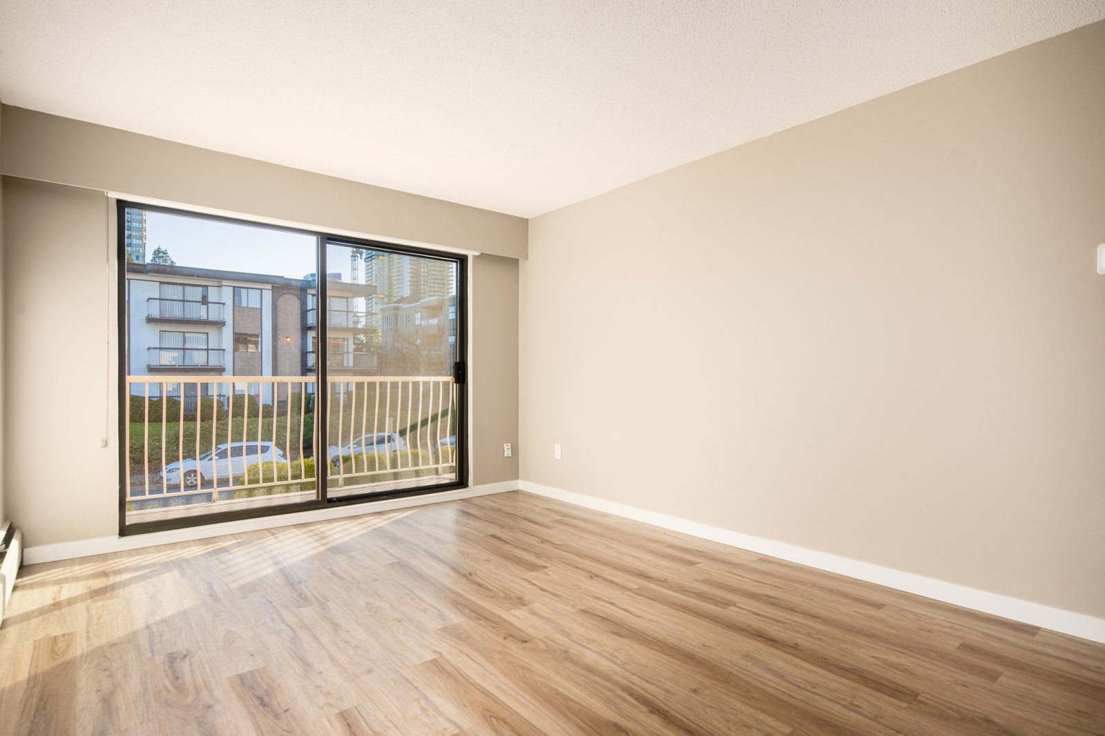 Empty room with light wood flooring, beige walls, and a large sliding glass door leading to a balcony overlooking an urban street and nearby buildings.