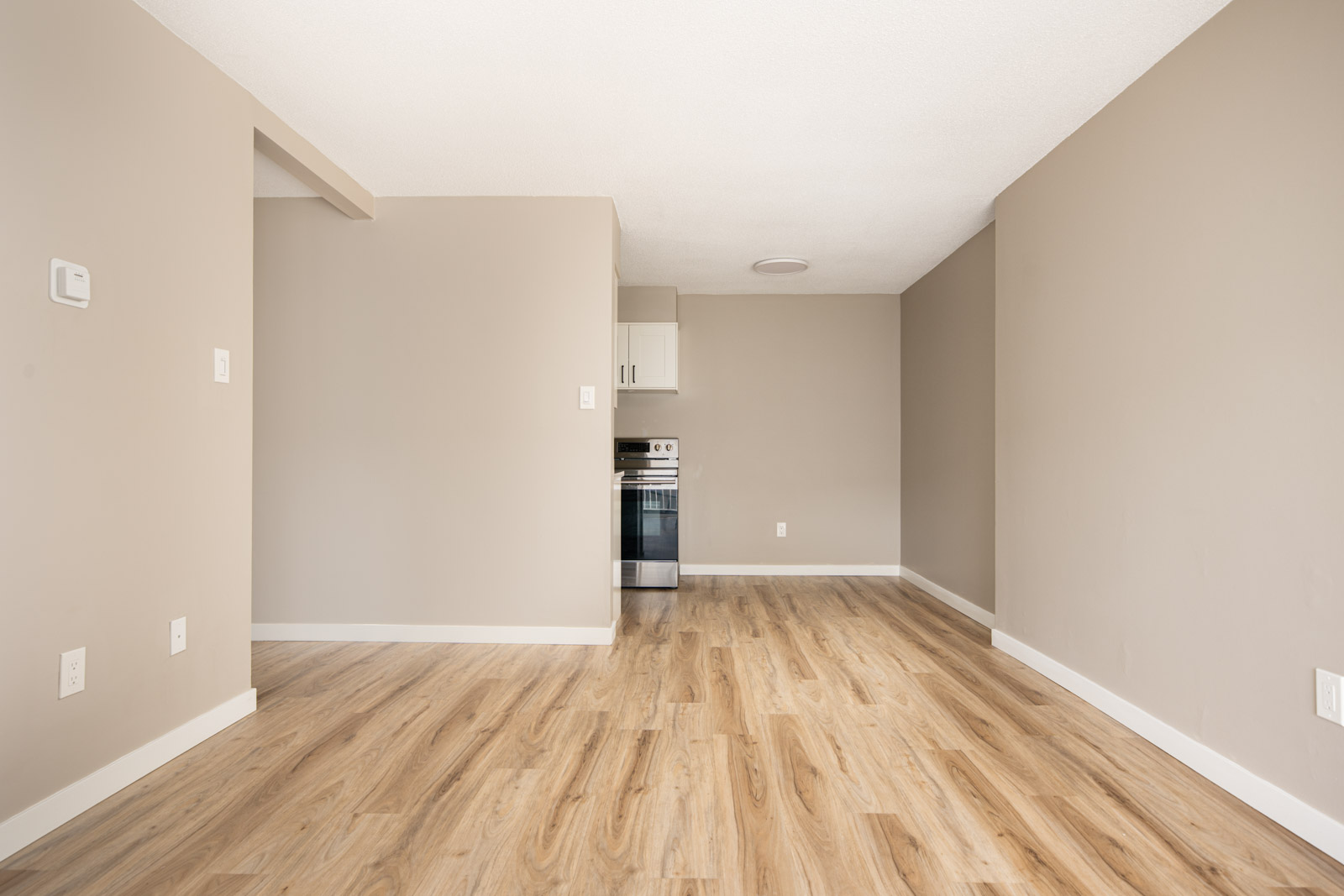 Empty room with light wood flooring, beige walls, and a small kitchen area at the back featuring a stainless steel oven.