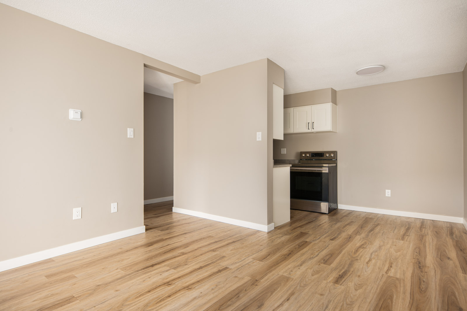 Empty room with light wood flooring, beige walls, and a small kitchen area with white cabinets and a stainless steel oven.
