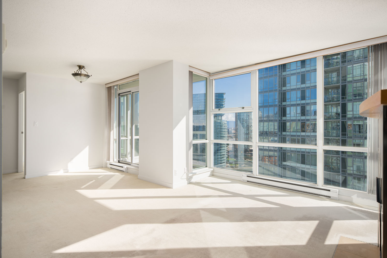 Bright, empty apartment living room with large windows, beige carpet, and cityscape views. Sunlight streams in, casting shadows across the floor.