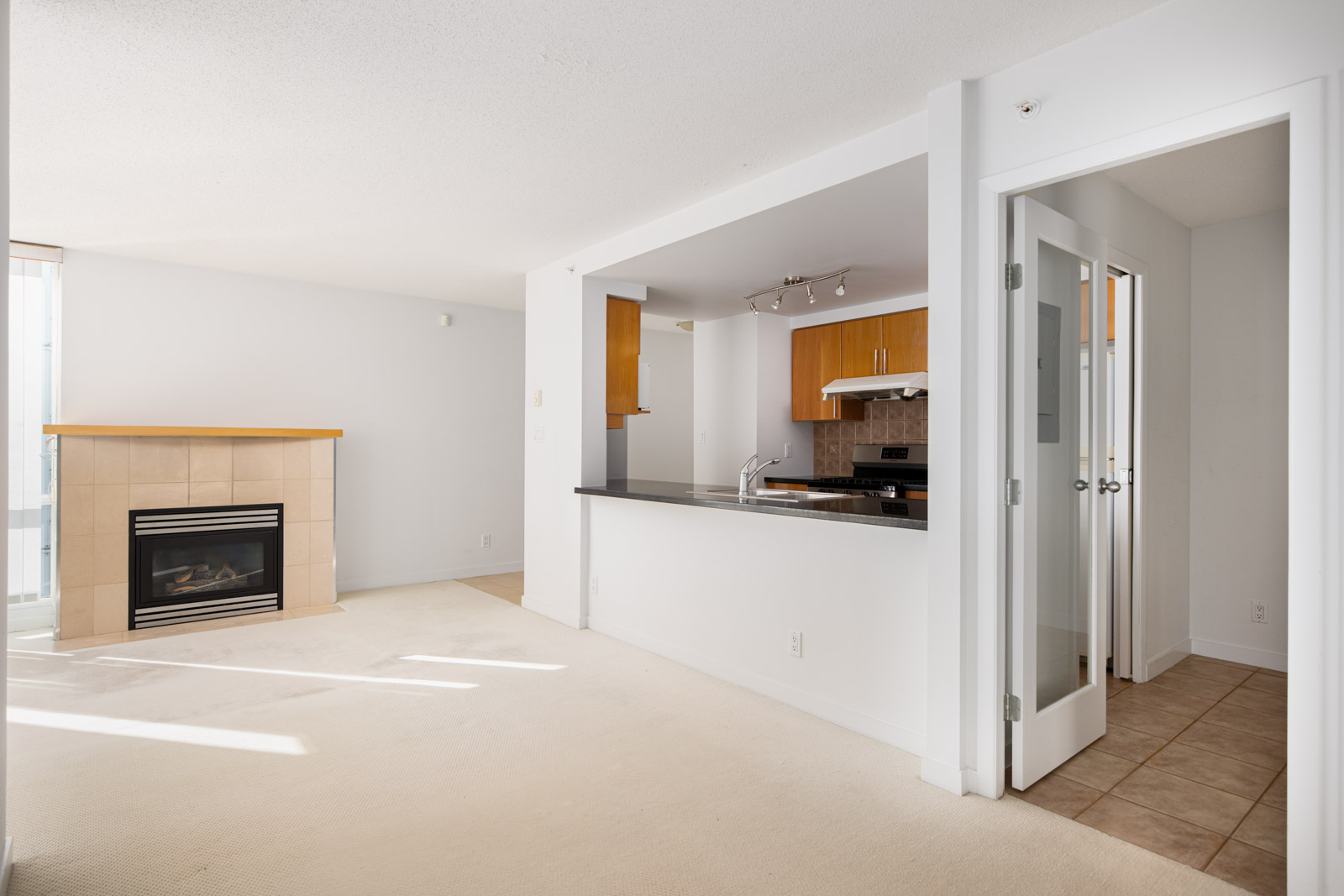 Unfurnished apartment with beige carpet, a fireplace, open kitchen with wood cabinets, and double glass doors leading to a tiled area.