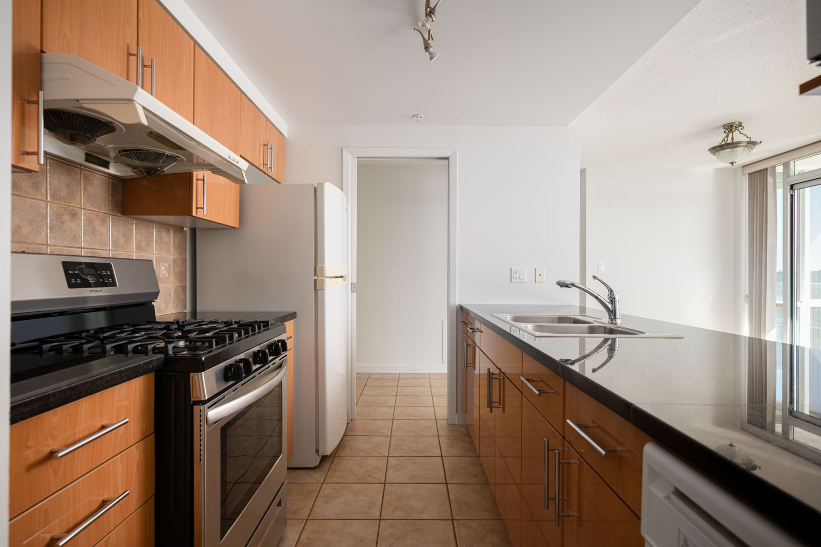 Modern kitchen with wood cabinets, stainless steel gas stove, white refrigerator, double sink, tiled floor, and a sliding glass door leading to a bright dining area.