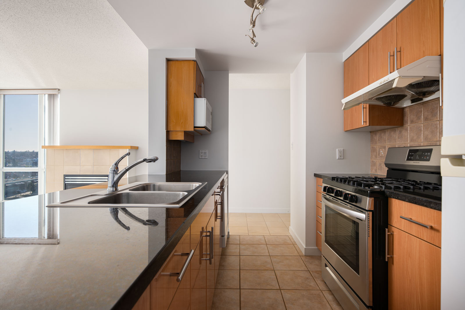Modern kitchen with wooden cabinets, black countertops, stainless steel appliances, double sink, tile flooring, and a window letting in natural light.