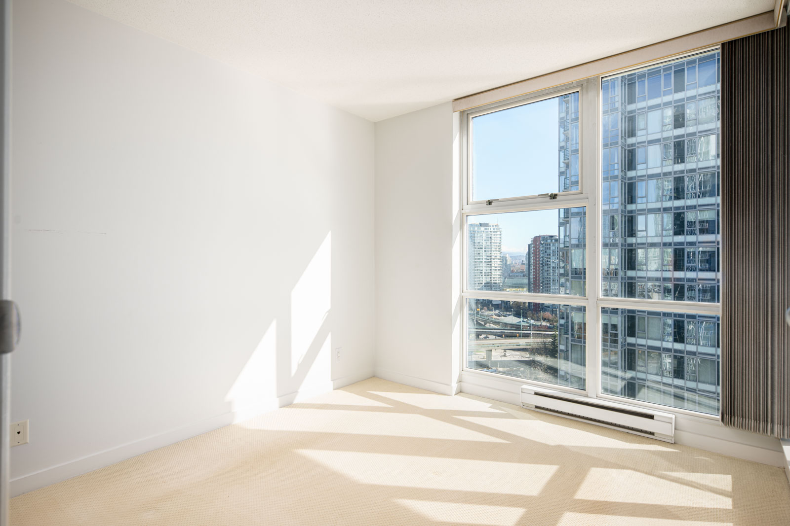 Bright, empty room with large window, beige carpet, and view of city buildings under clear sky. Sunlight casts shadows on the walls and floor.