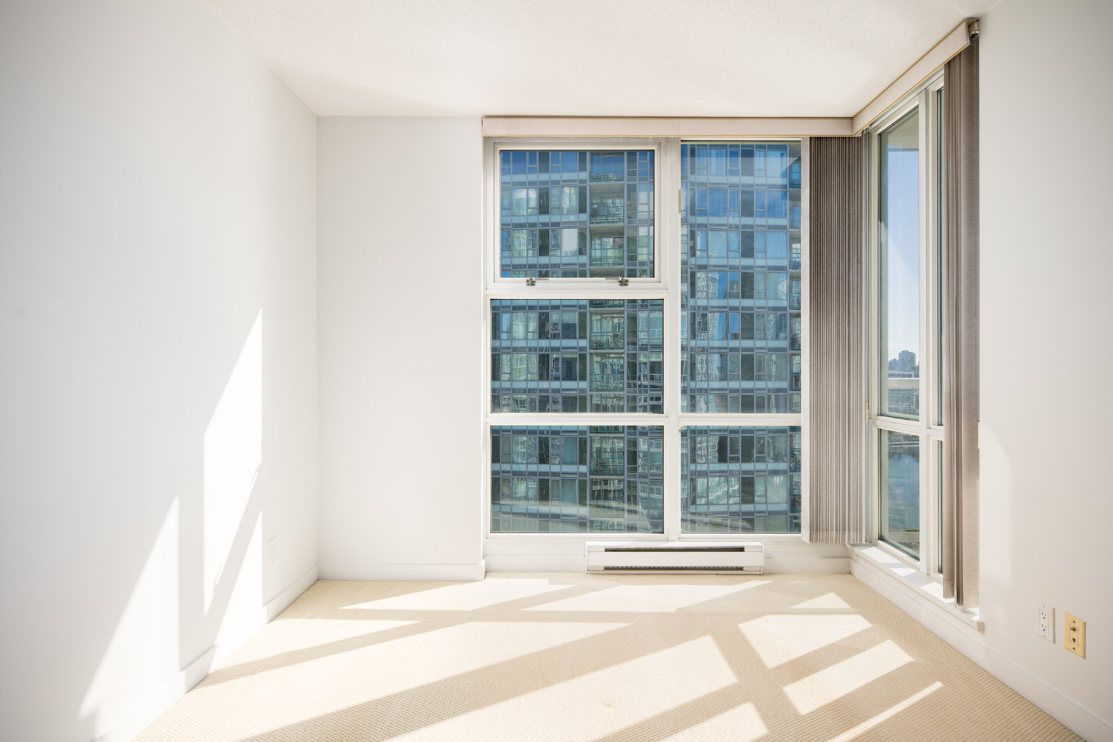 Empty room with beige carpet, large floor-to-ceiling windows, vertical blinds, and a view of modern glass apartment buildings outside.