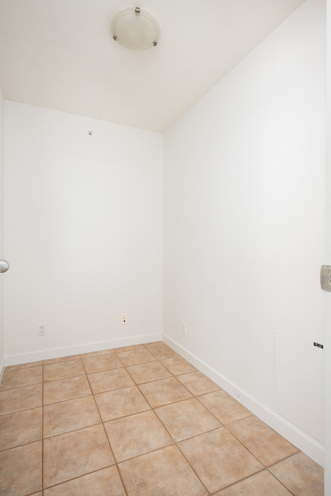 Empty room with white walls, a ceiling light fixture, beige floor tiles, electrical outlets, and no furniture or decor.