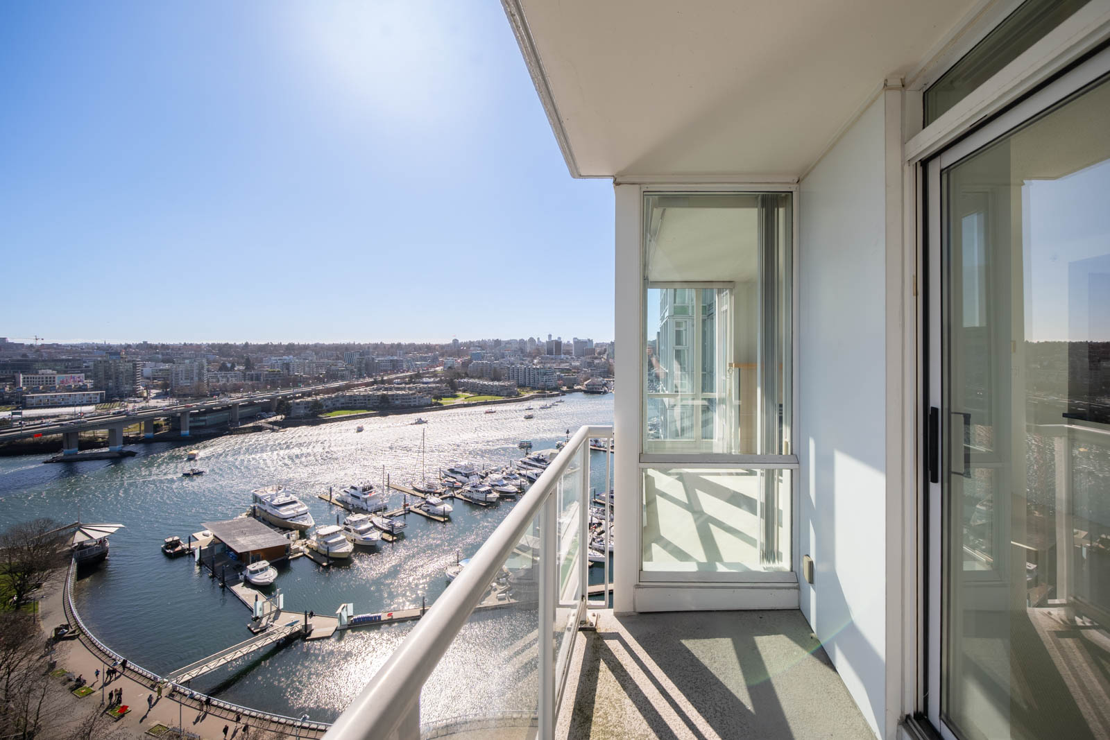 High-rise balcony overlooking a marina with several docked boats, a bridge, and a cityscape under a clear blue sky.