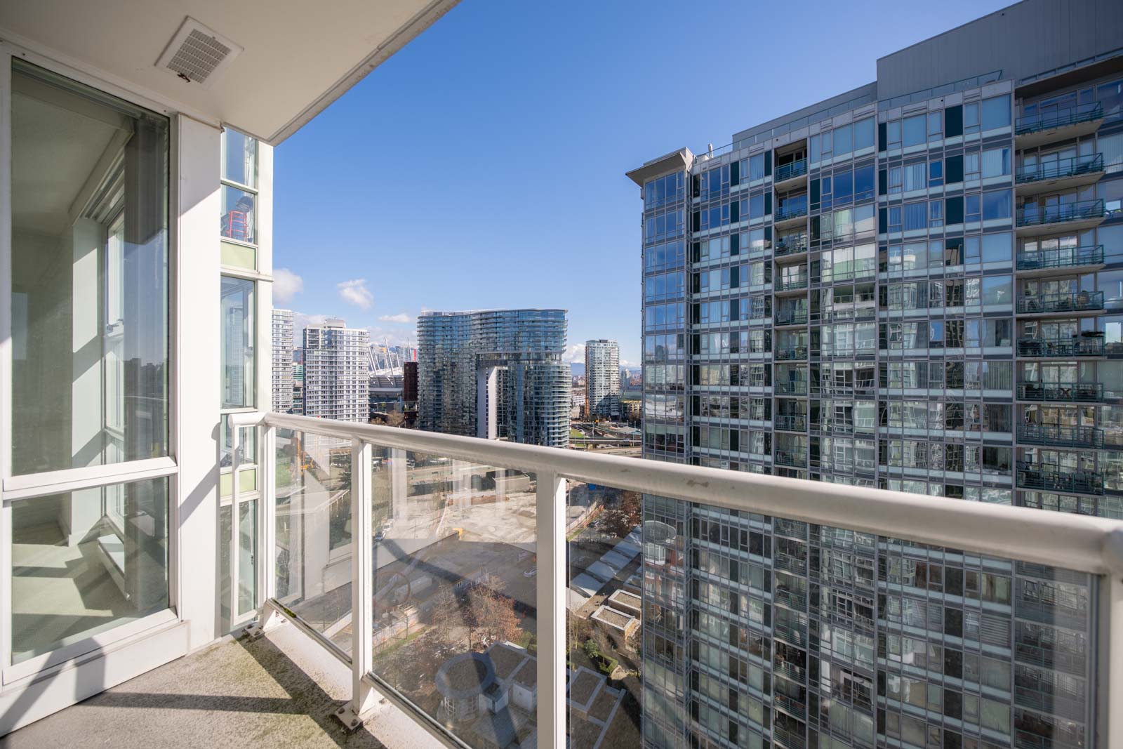 A high-rise apartment balcony with glass railing overlooks modern glass buildings and a cityscape under a clear blue sky.
