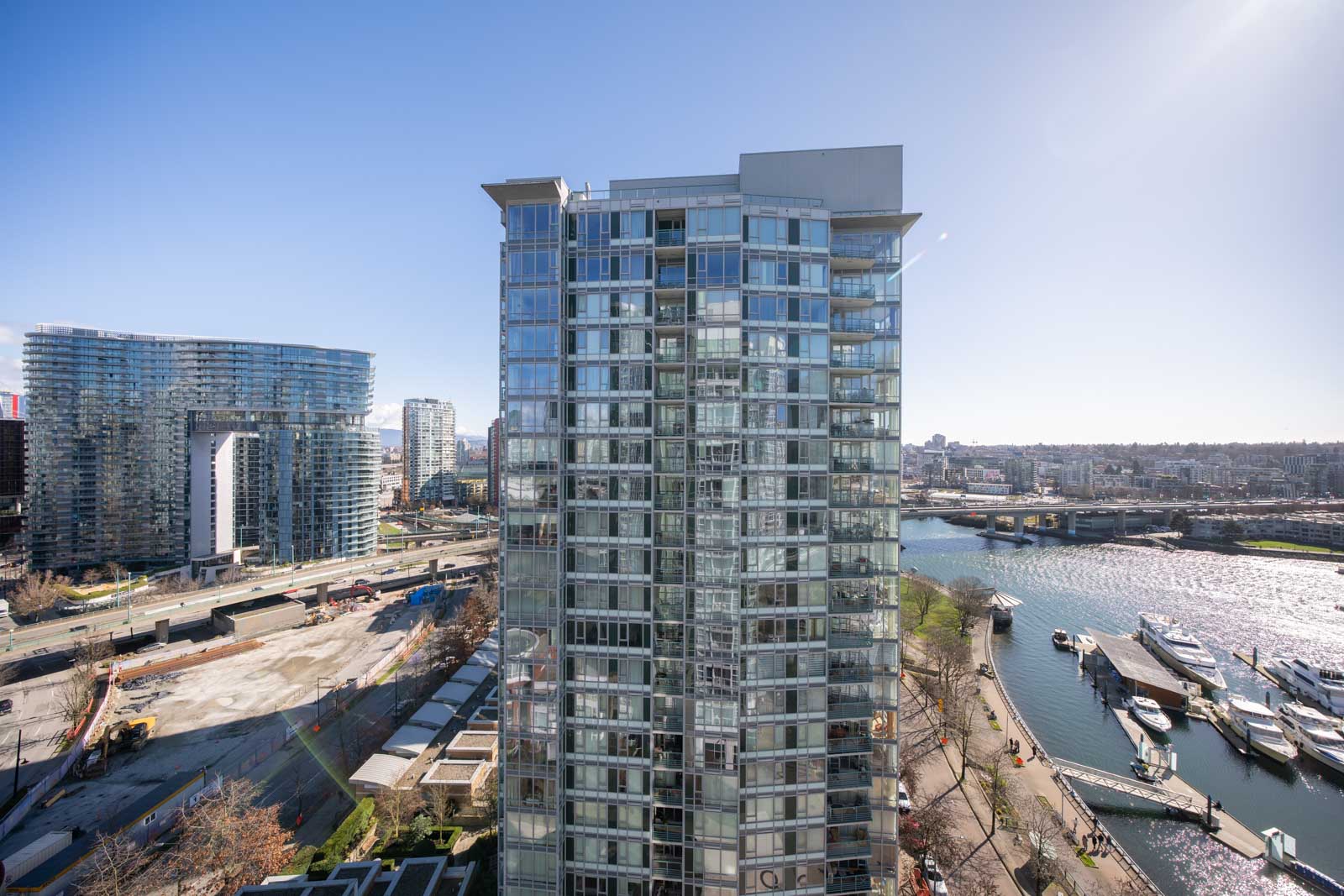 High-rise glass apartment building with balconies near a river, adjacent to a marina and construction site, under a clear blue sky.