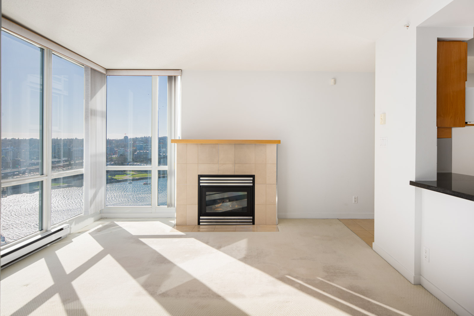 Bright living room with large windows, a gas fireplace with tile surround, light carpet flooring, and partial view of a kitchen area to the right.