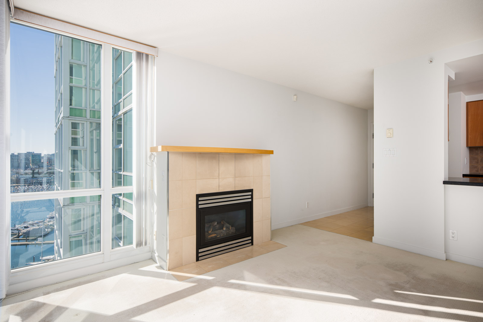 Bright living room with a gas fireplace, large windows overlooking water and city buildings, and light-colored carpet and walls.