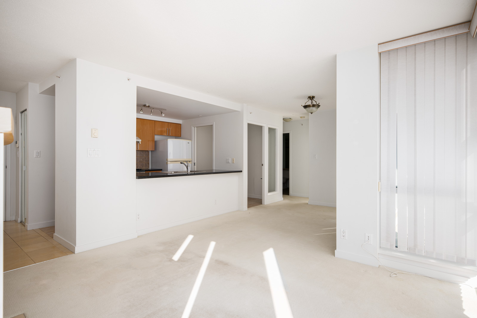 Unfurnished apartment interior with white walls, open kitchen, carpeted living area, and vertical blinds covering large windows letting in natural light.
