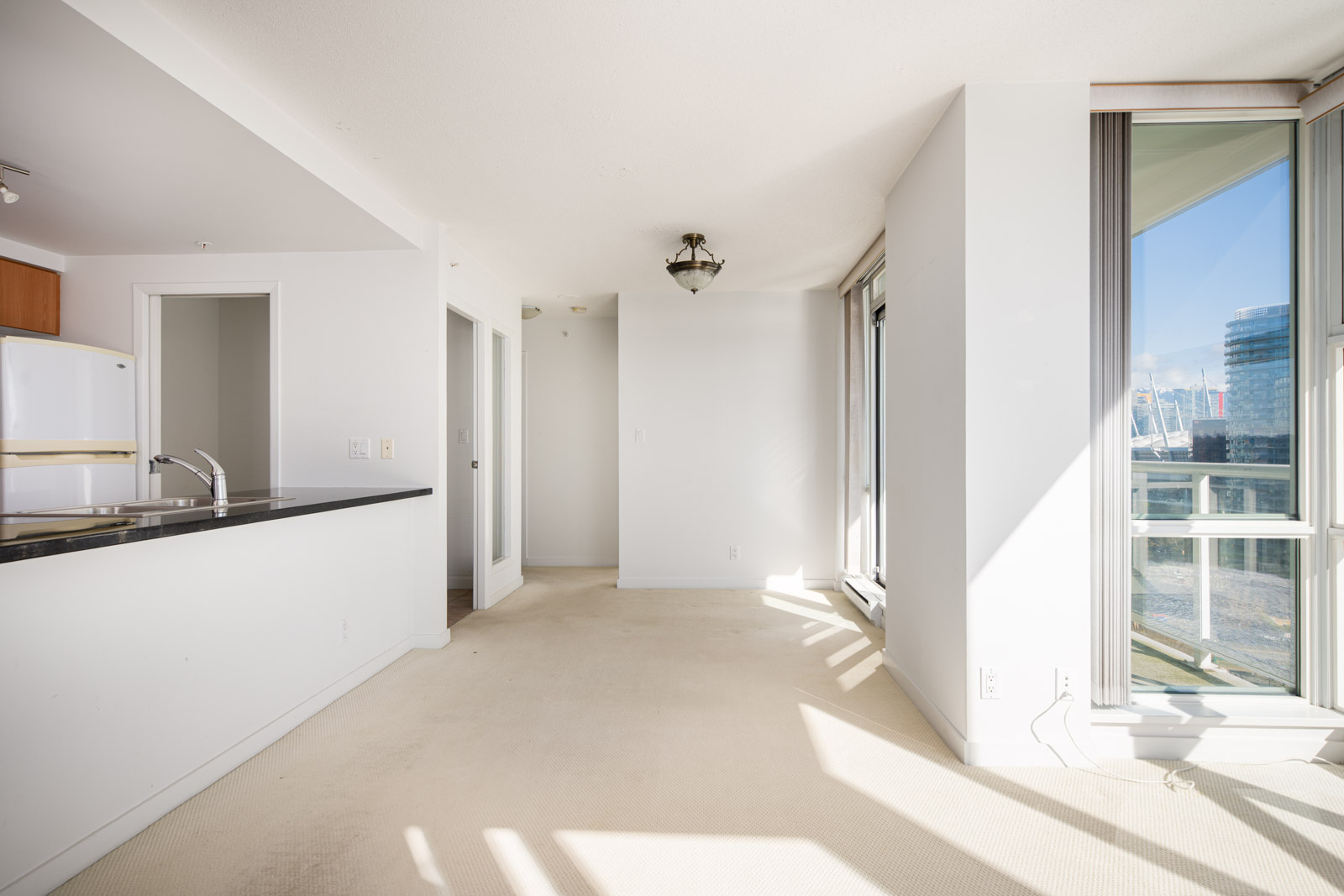 Bright, empty apartment interior with beige carpet, white walls, a kitchen with black countertops, large windows, and a ceiling light fixture.