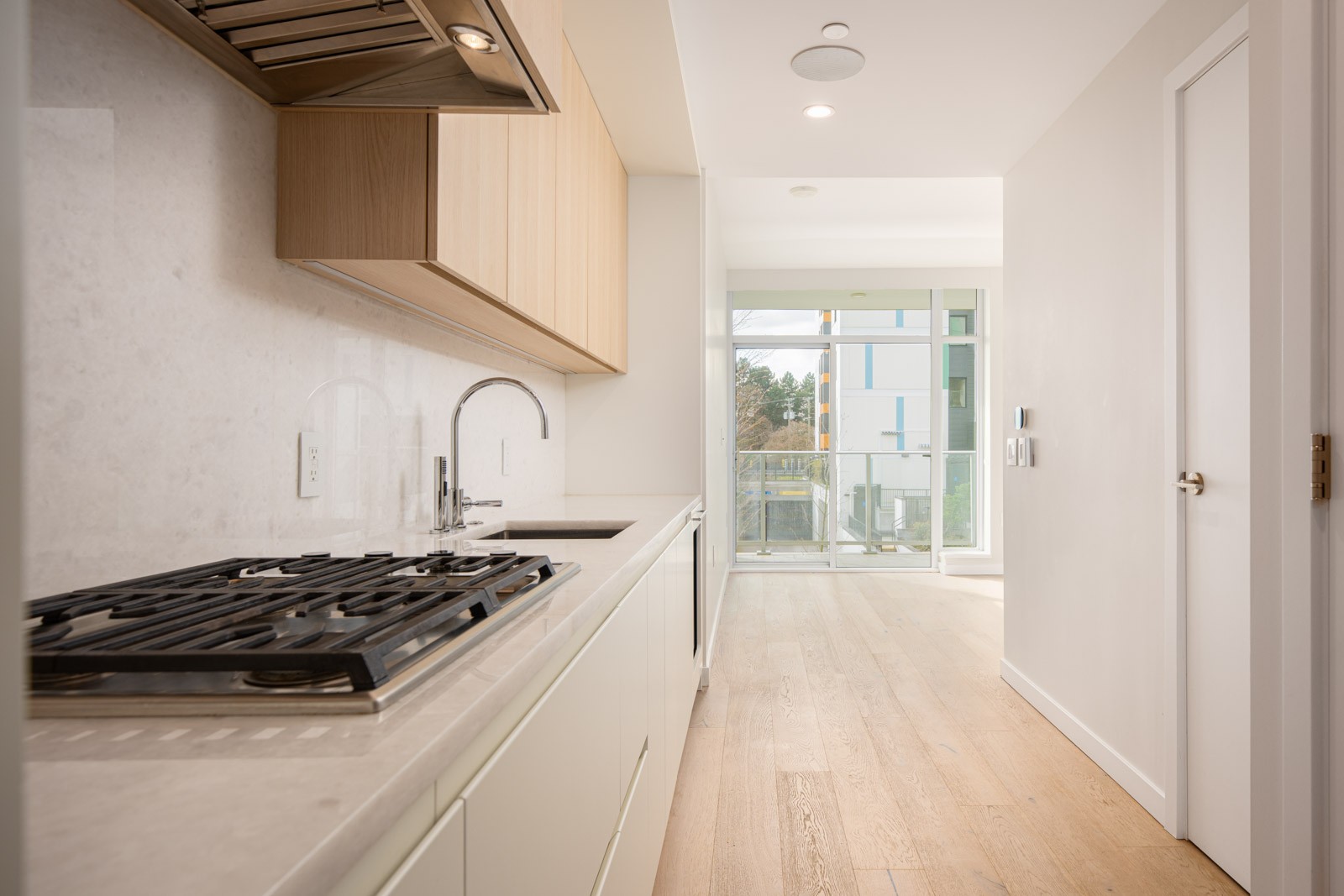 Modern kitchen with a gas cooktop, light wood cabinets, white countertops, and a sink, leading to a bright living area with large windows and a balcony.