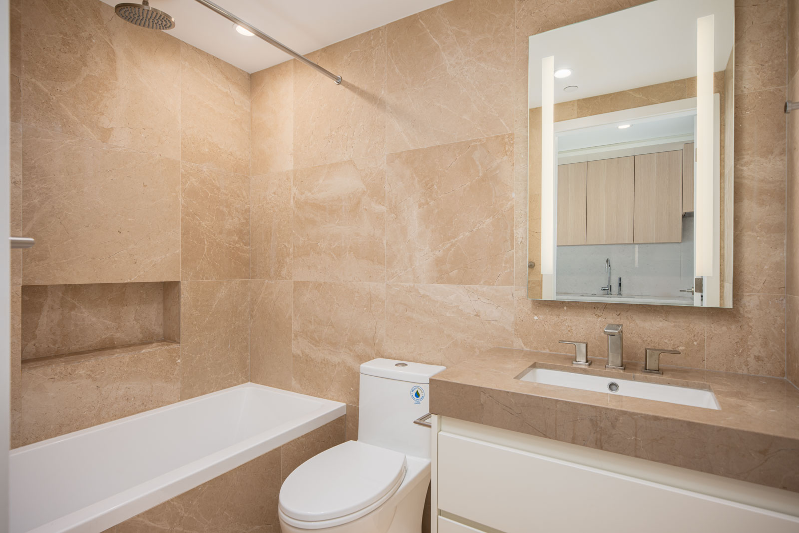 Modern bathroom with beige marble tiles, a built-in bathtub with shower, a toilet, a sink with a large mirror, and minimalist fixtures.