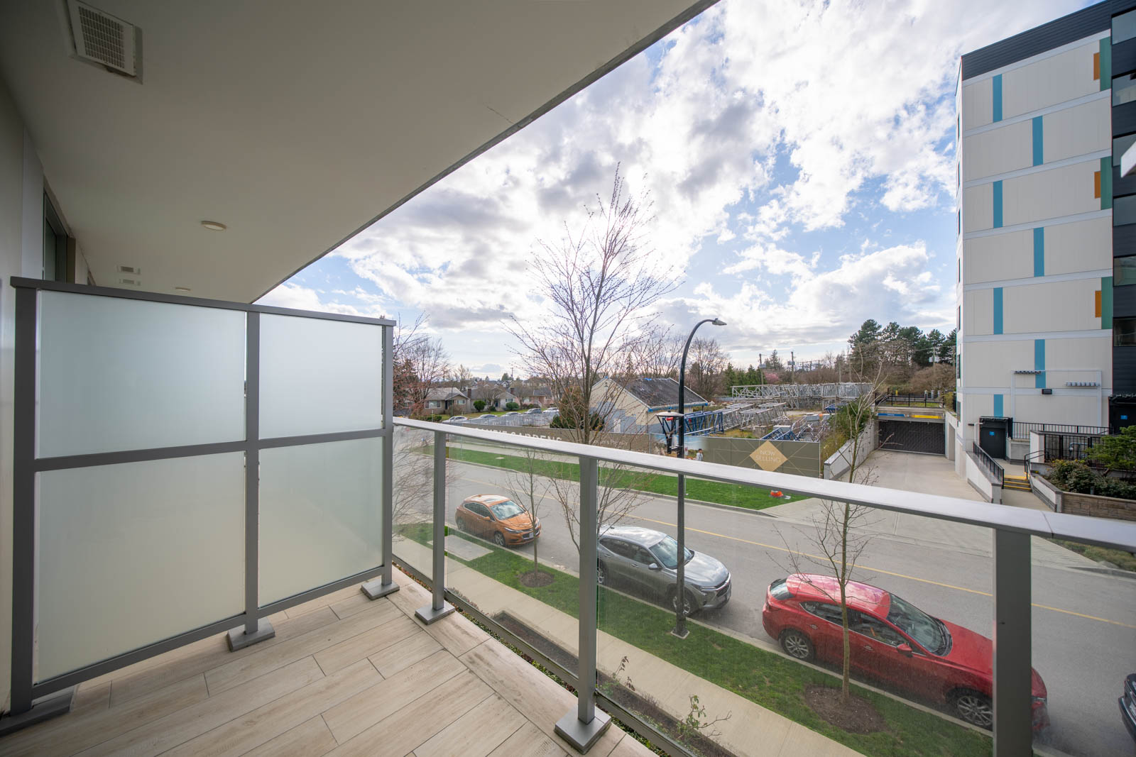 View from a balcony with glass railing and frosted privacy panel, overlooking a street with parked cars, trees, and a modern building under a partly cloudy sky.