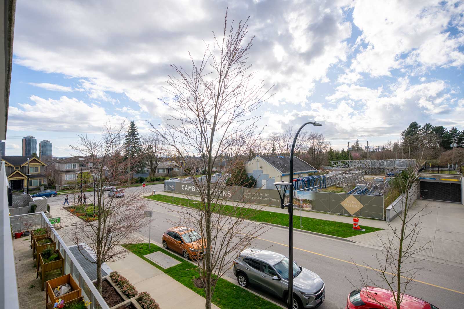 Street view featuring parked cars, residential houses, leafless trees, and a fenced construction area under a partly cloudy sky.