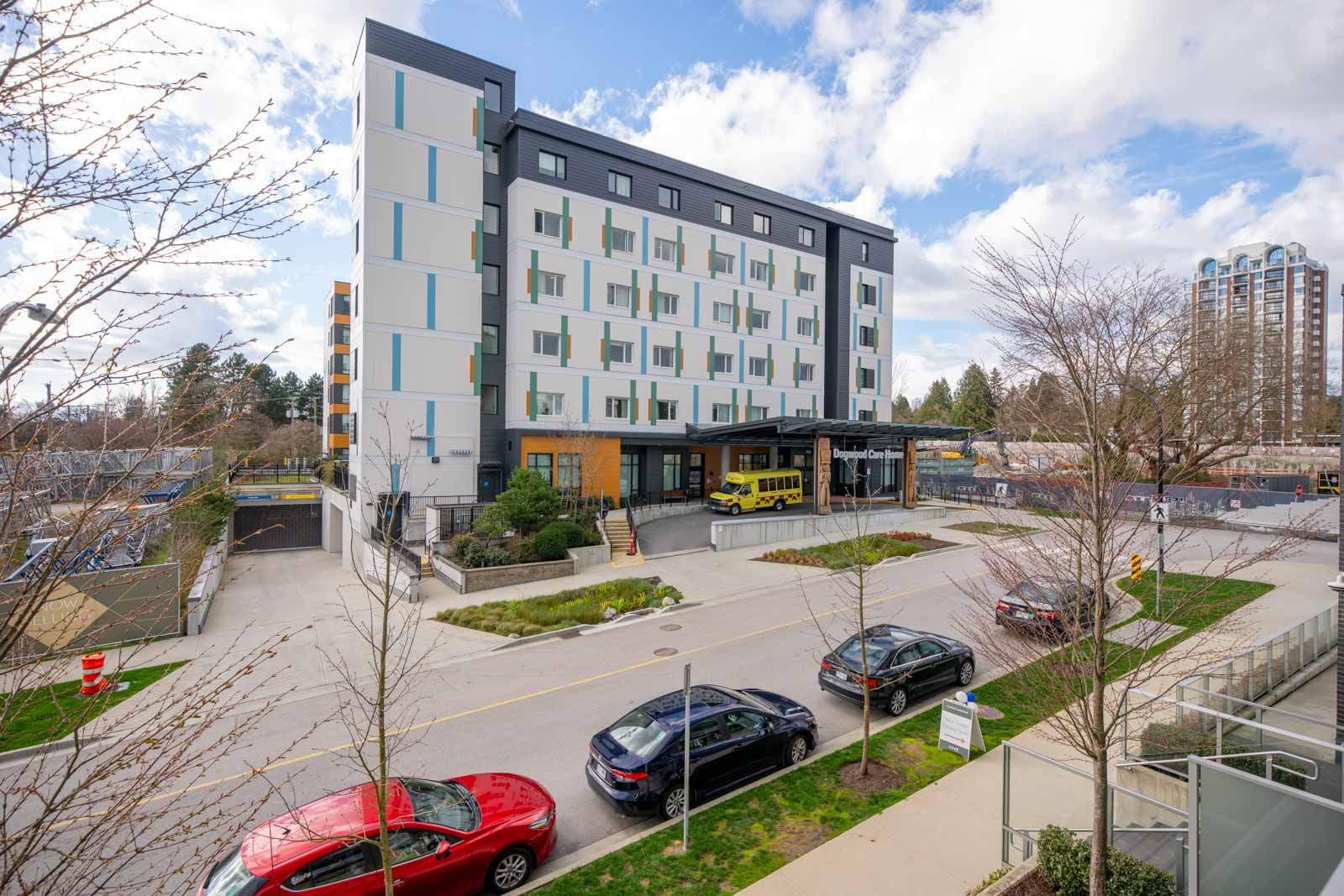 A modern six-story building with a covered entrance, several parked cars, and a yellow school bus in front, set against a partly cloudy sky.