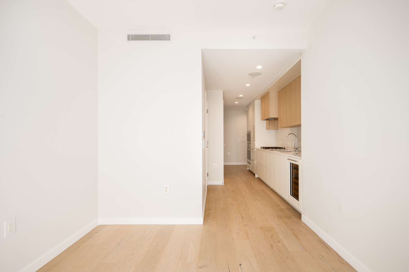 A narrow modern kitchen with light wood floors, white walls, built-in light wood cabinets, and recessed lighting in the ceiling.