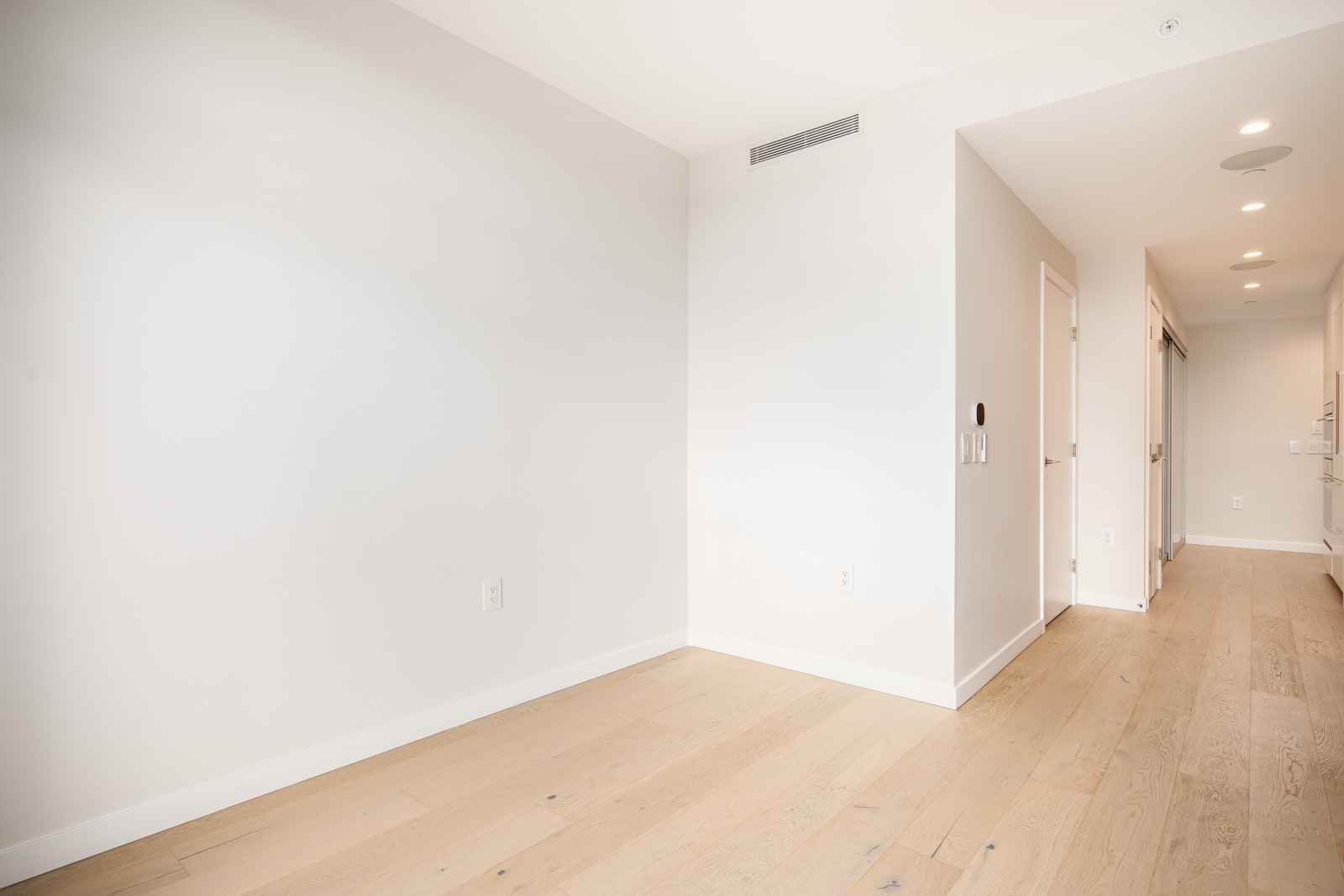 Bright, empty room with light wooden floors, white walls, and recessed lighting, leading to a hallway with doors and a visible kitchen area at the far end.