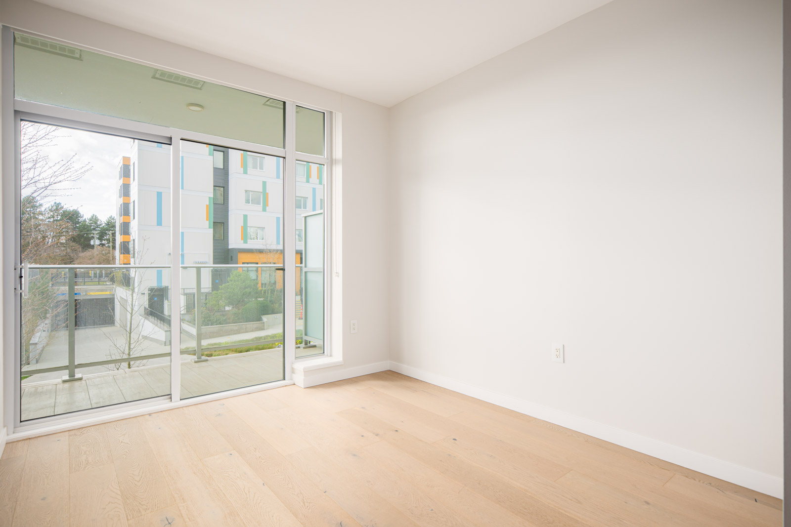 Empty room with light wood flooring, white walls, and large glass sliding doors leading to a balcony with a view of a modern apartment building.