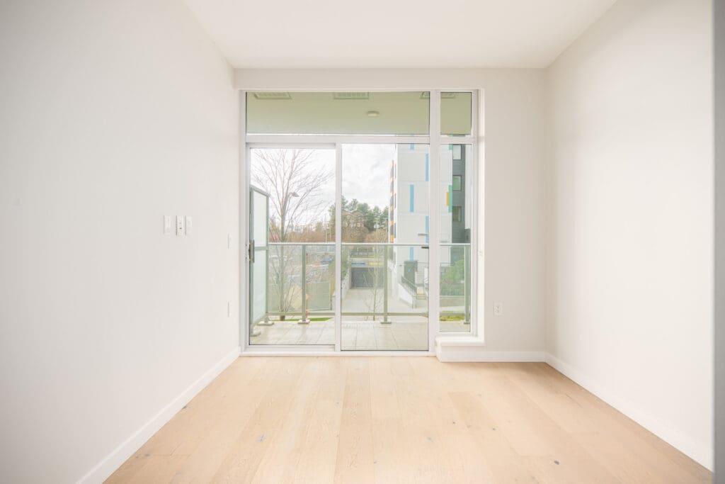 Empty room with light wood flooring, white walls, and large sliding glass doors leading to a balcony with a view of trees and a modern building.