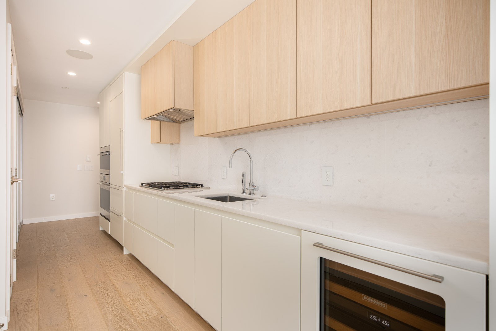 A modern kitchen with light wood cabinets, white countertops, built-in appliances, a sink, and a wine fridge, featuring minimalist design and natural wood flooring.