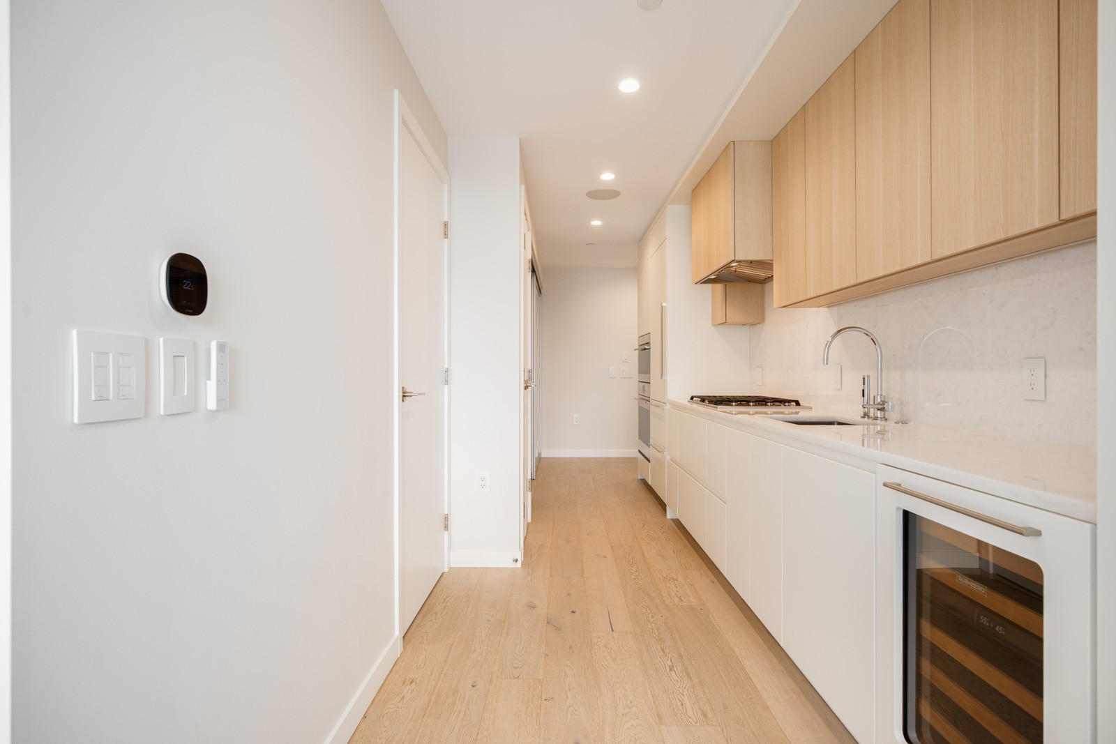 Modern galley kitchen with light wood flooring, white cabinets, wood-accented upper cabinets, built-in appliances, and a wine fridge along one wall.