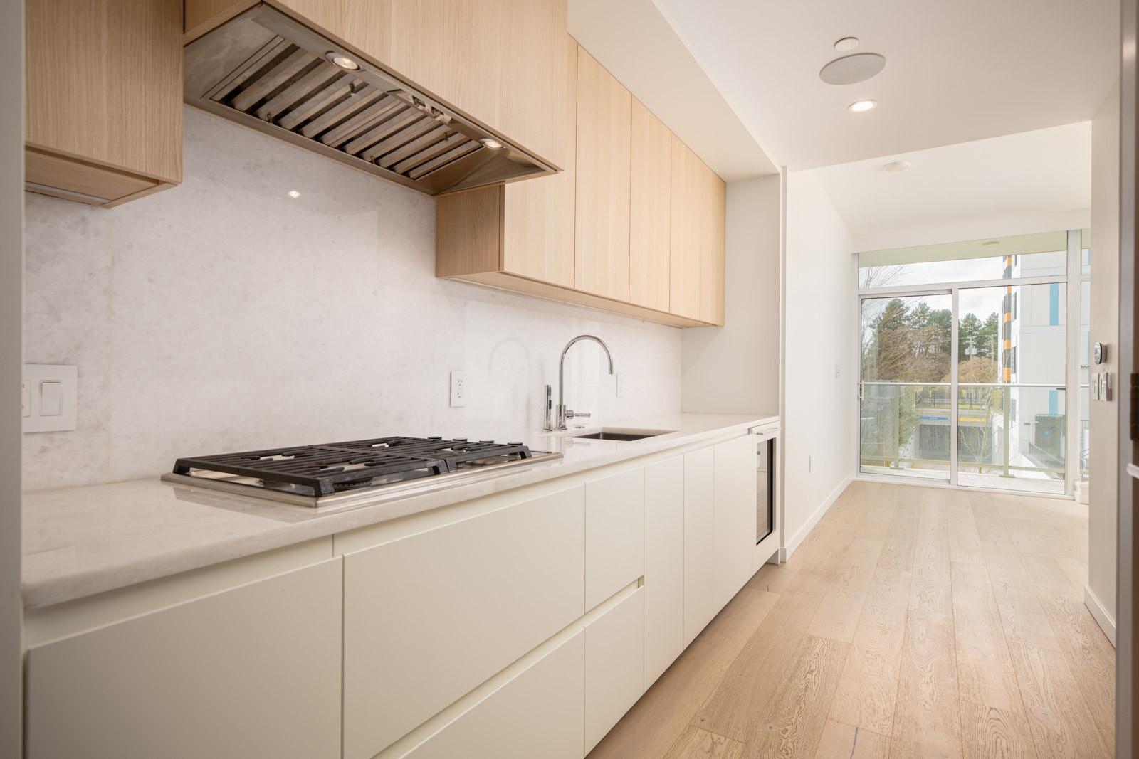 Modern galley kitchen with light wood cabinets, white countertops, gas stove, built-in oven, and a large window leading to a balcony with views outside.