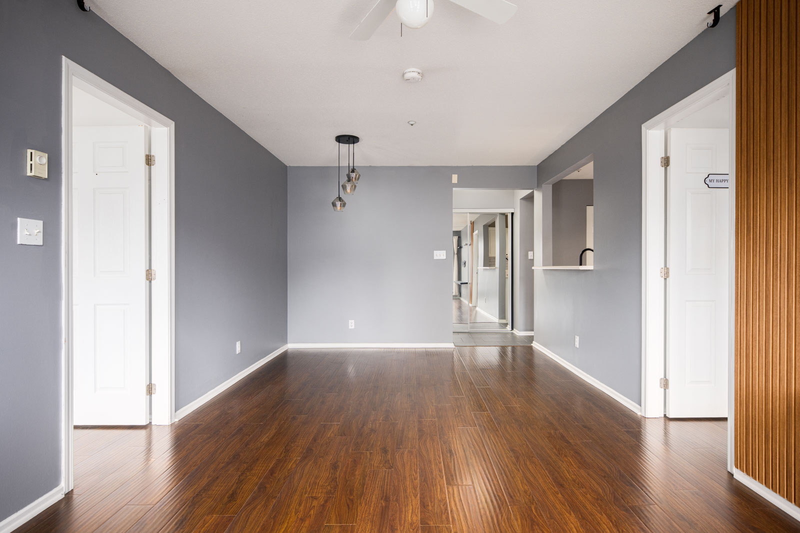 Empty room with wood flooring, gray walls, ceiling fan, pendant lights, and several open doorways leading to adjacent rooms and a hallway.
